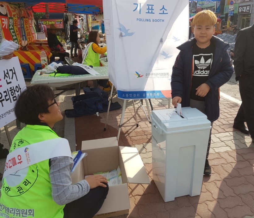2018년 3월 30일 칠원읍사무소 앞 도로변 삼칠민속줄다리기 행사에서 홍보주임 및 지원단이 공명선거 행사 중 사전투표 체험 중 투표한 어린이를 대상으로 사진을 촬영하였습니다.
